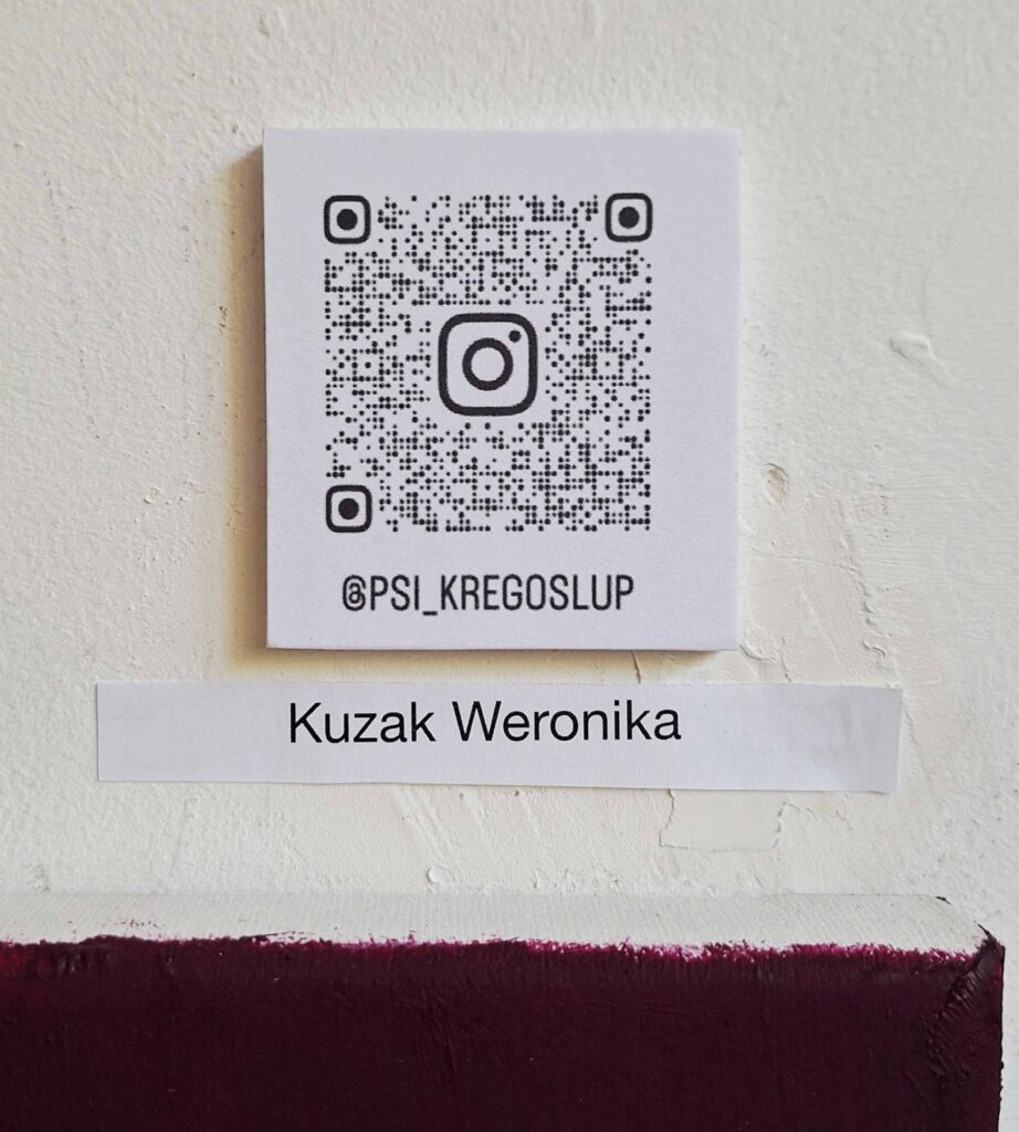 Tabliczka z nazwiskiem Weroniki Kuzak oraz kodem QR prowadzącym do profilu artystki na Instagramie podczas wystawy końcoworocznej ASP 2026
